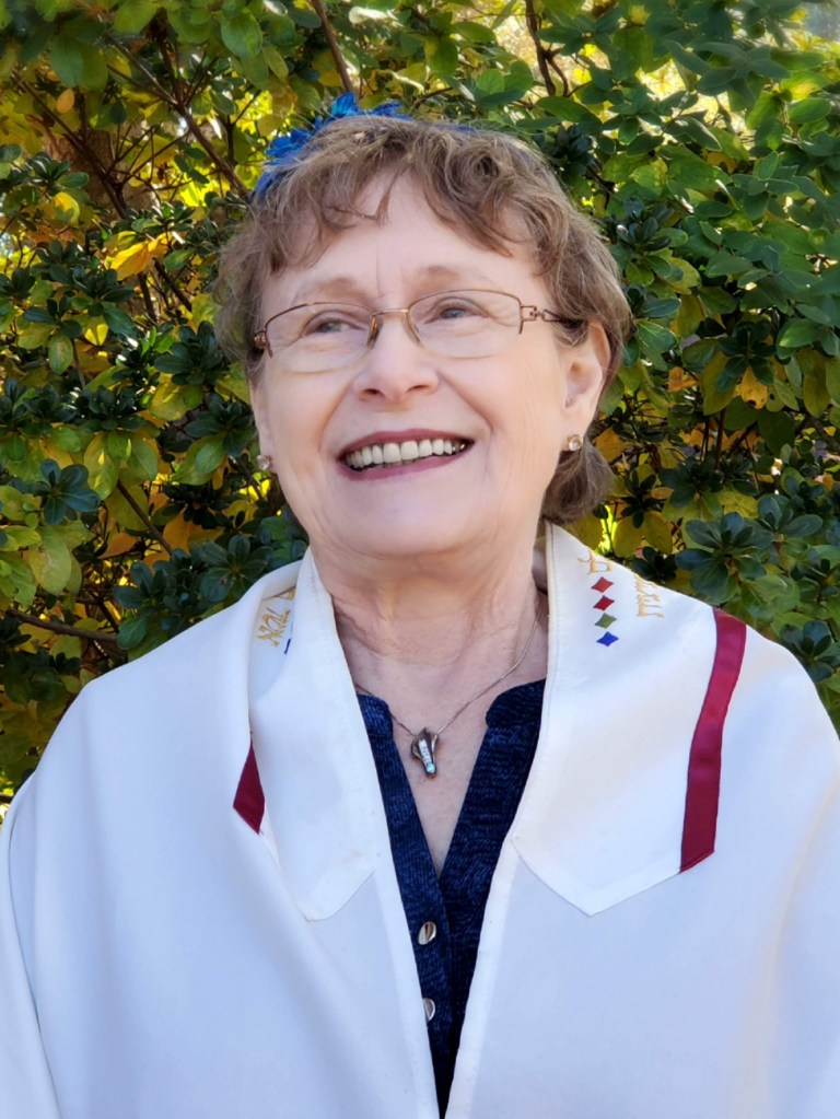 Photo of a Reb Nancy, an ordained Jewish Rabbinic Pastor, wearing a white tallit and looking into the distance.